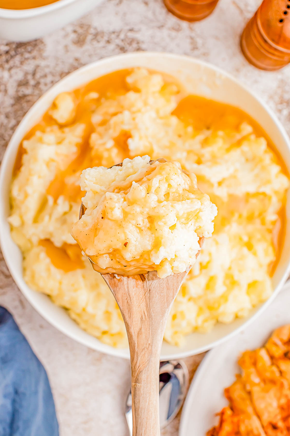 A wooden spoon holding creamy mashed potatoes with gravy above a bowl of the same dish on a table.