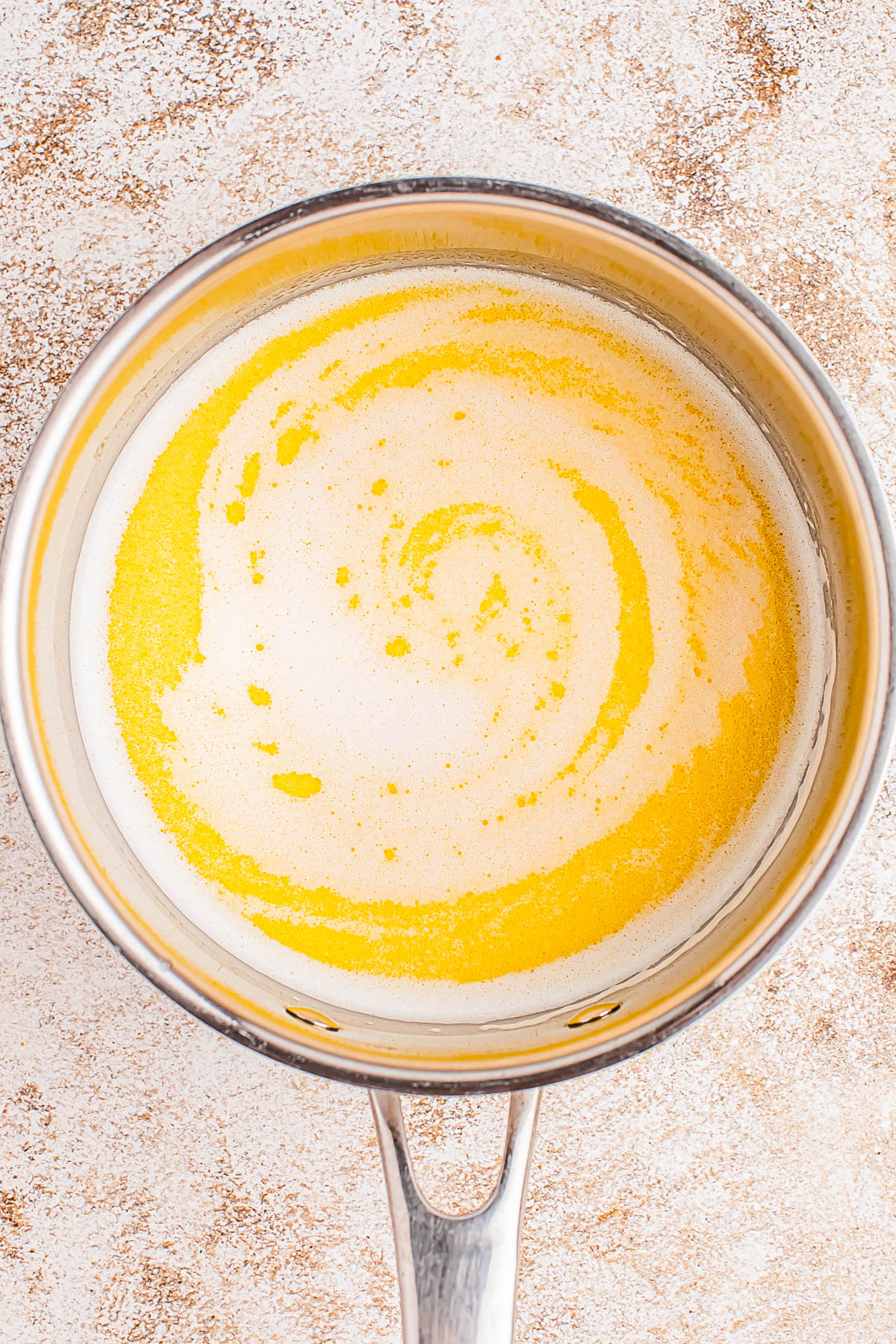 A saucepan containing melted butter with a swirling pattern on the surface, placed on a textured countertop.