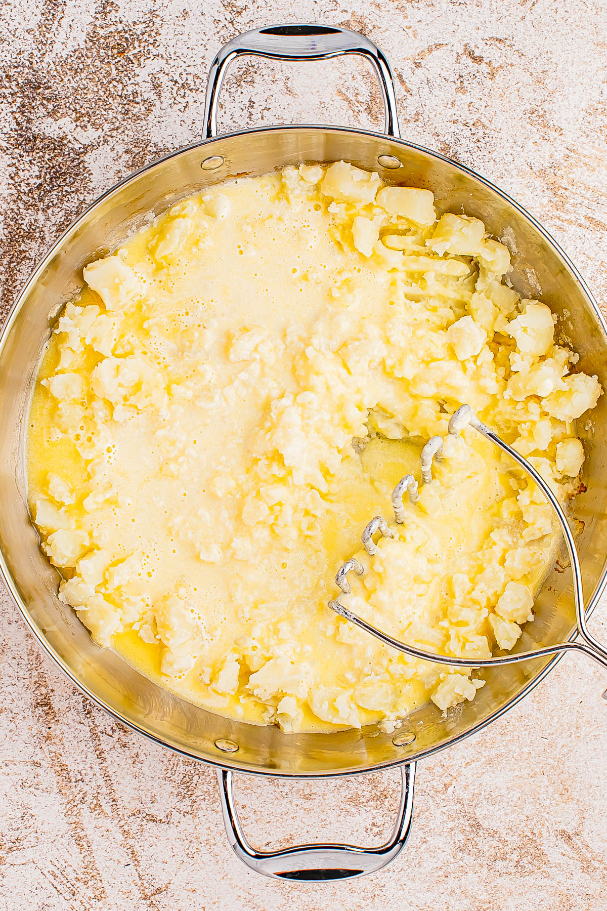 Mashed potatoes in a metallic pot with a potato masher, showing a creamy texture and buttery appearance.