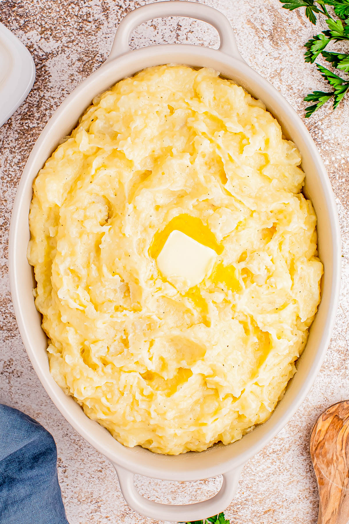 Bowl of mashed potatoes topped with a pat of butter, on a textured surface with parsley nearby.