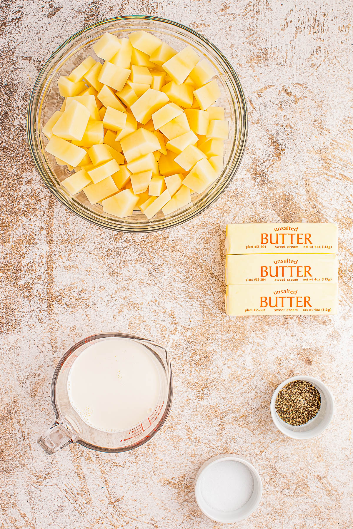 Ingredients for mashed potatoes: a bowl of cubed potatoes, three sticks of unsalted butter, a measuring cup of cream, a small dish of salt, and another of pepper on a textured surface.