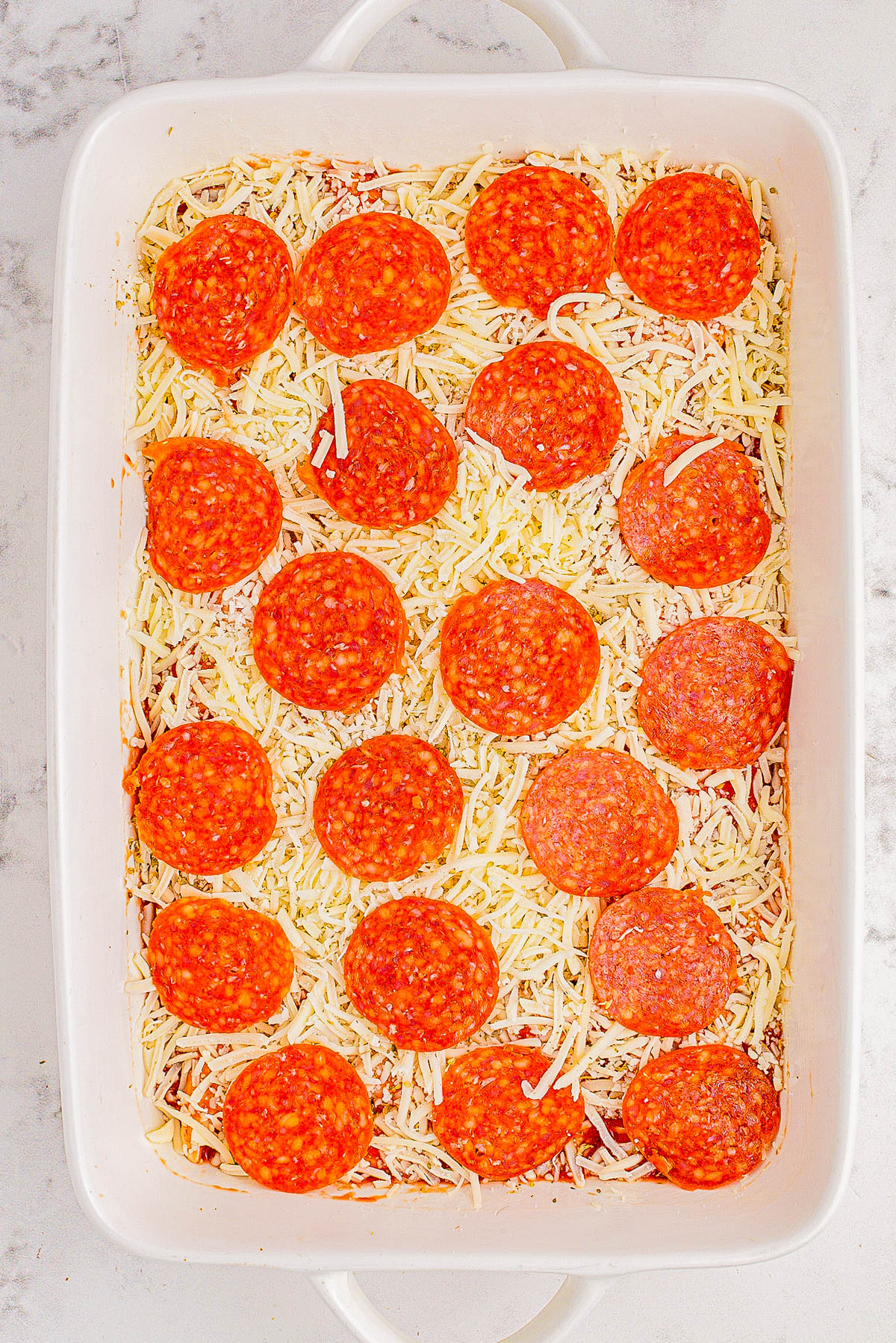 A baking dish filled with shredded cheese and topped with slices of pepperoni.