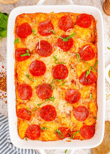 A baked pepperoni pizza casserole in a rectangular dish, topped with melted cheese and fresh basil.