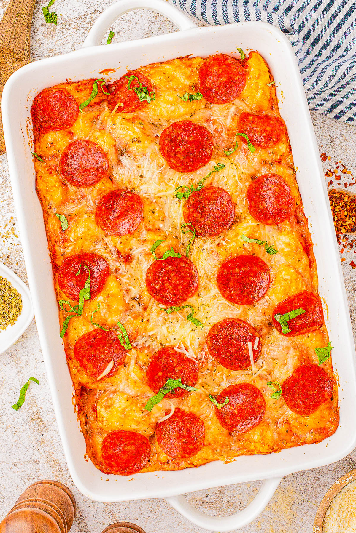 A rectangular baking dish filled with baked pepperoni pizza casserole topped with sliced pepperoni and garnished with herbs.