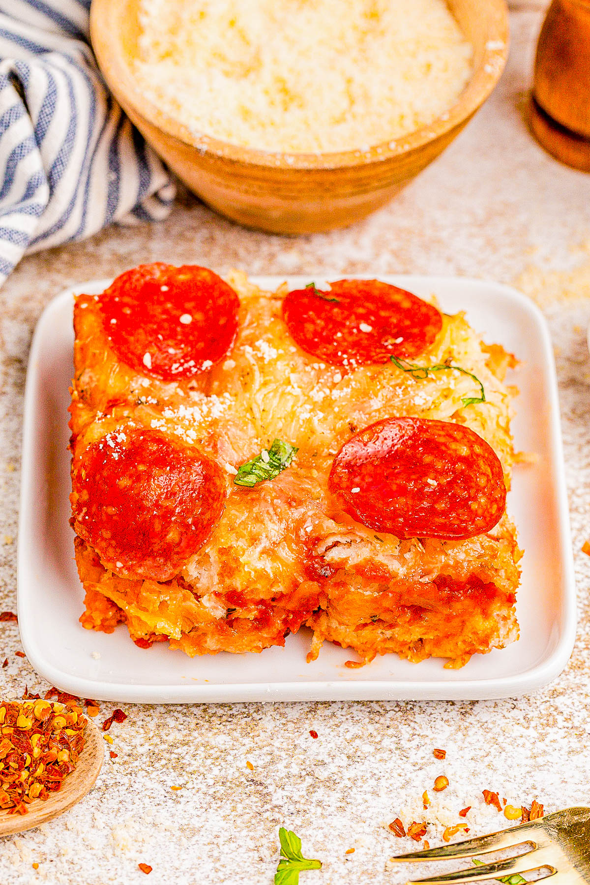 A slice of pizza lasagna topped with pepperoni and cheese on a white square plate, sprinkled with herbs. A bowl of Parmesan and a striped napkin are in the background.