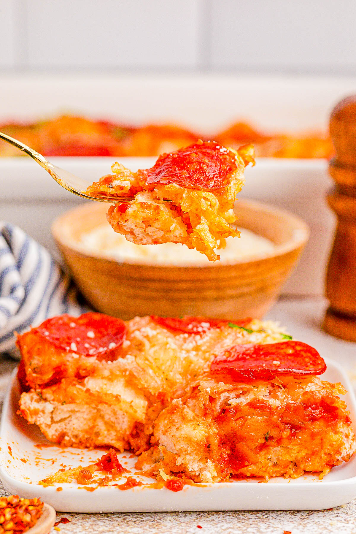 A fork holds a bite of a pepperoni pizza casserole over a plate. The dish features layers of cheese, sauce, and pepperoni slices. A bowl of grated cheese is in the background.