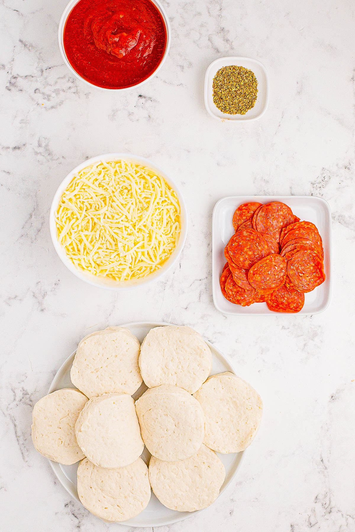 Ingredients for mini pizzas arranged on a marble surface: tomato sauce, shredded cheese, pepperoni slices, Italian seasoning, and uncooked pizza dough rounds.