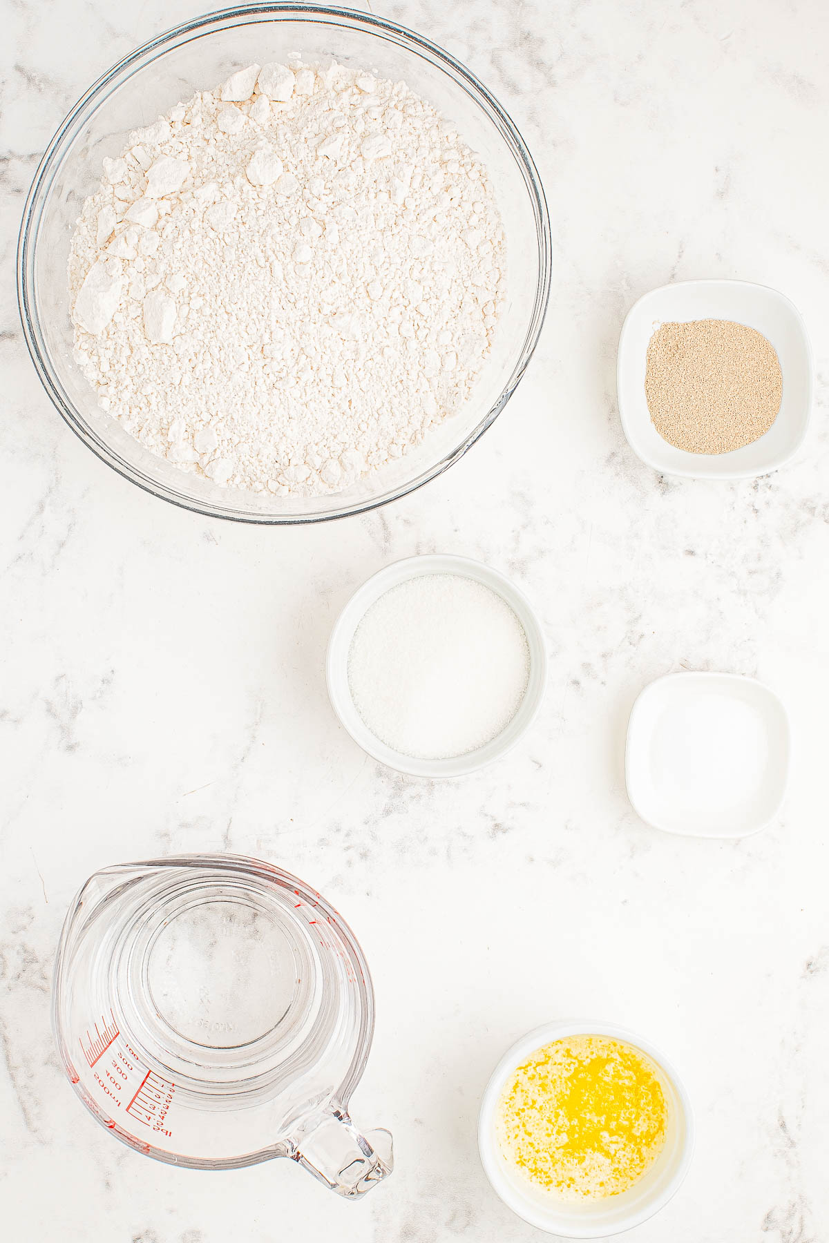 Baking ingredients on a marble surface: a bowl of flour, a small bowl of yeast, a bowl of sugar, a bowl of butter, and a measuring cup with water.