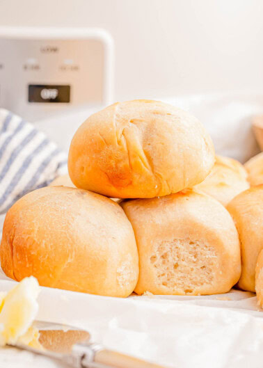 A stack of freshly baked dinner rolls on a table with a knife and butter in the foreground.