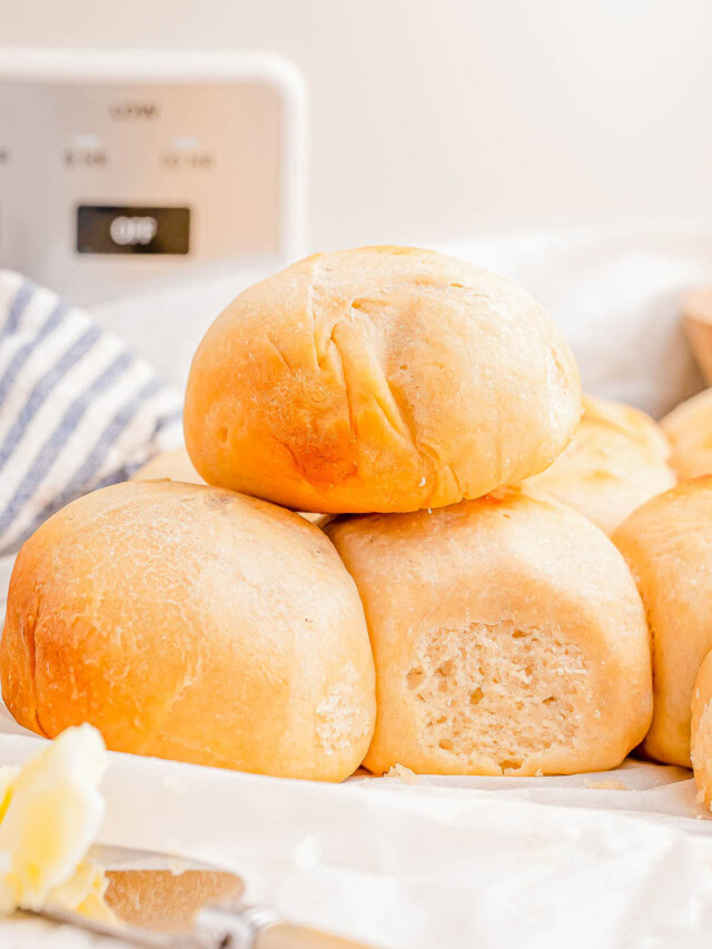 A stack of freshly baked dinner rolls on a table with a knife and butter in the foreground.