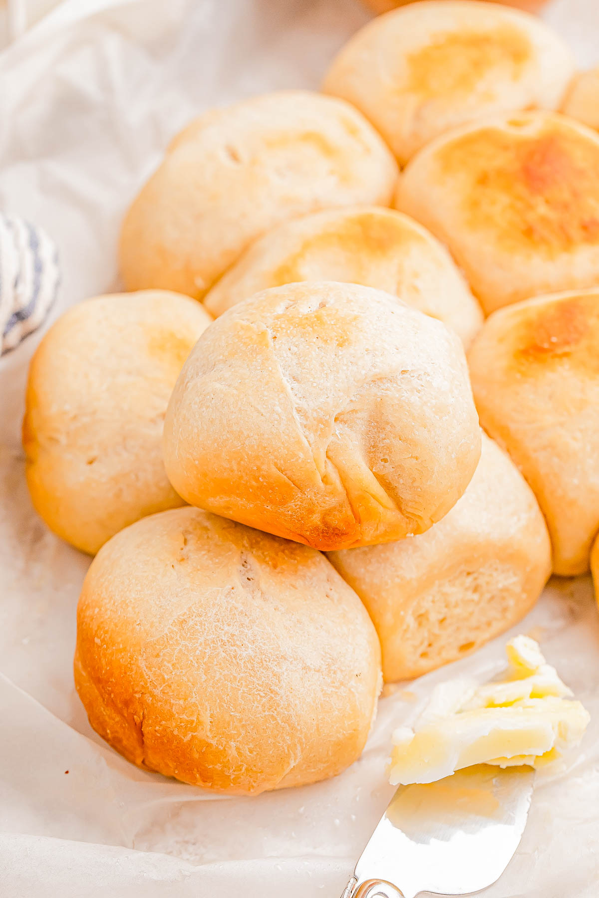 A stack of freshly baked bread rolls on parchment paper, with a buttered knife beside them.