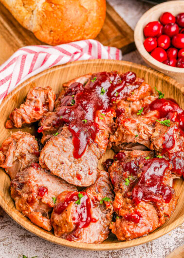 Sliced pork tenderloin topped with cranberry sauce in a wooden bowl, accompanied by a bowl of cranberries and a loaf of bread.