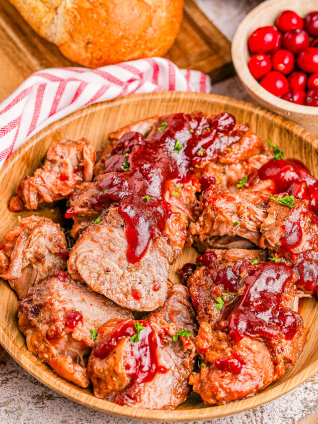 Sliced pork tenderloin topped with cranberry sauce in a wooden bowl, accompanied by a bowl of cranberries and a loaf of bread.