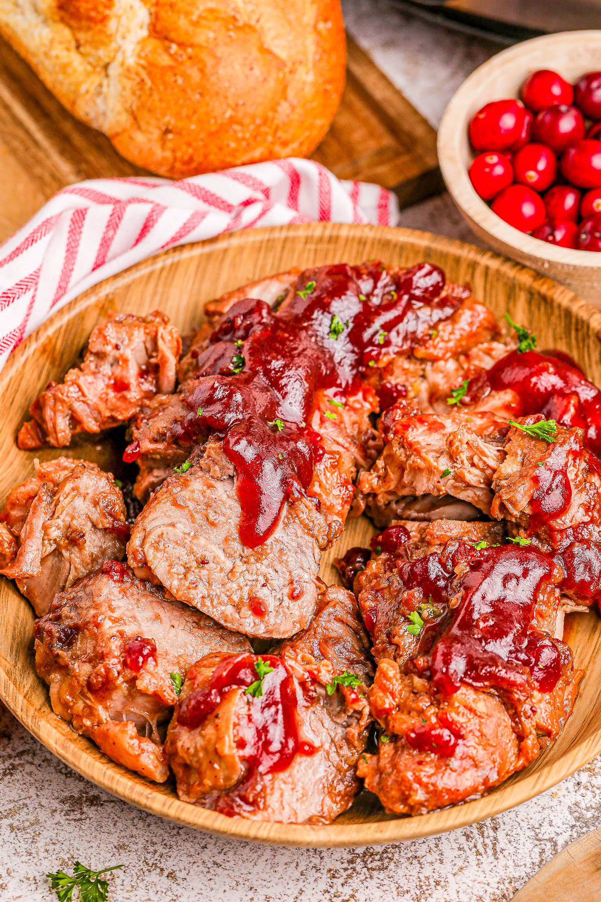 Sliced pork tenderloin topped with cranberry sauce in a wooden bowl, accompanied by a bowl of cranberries and a loaf of bread.