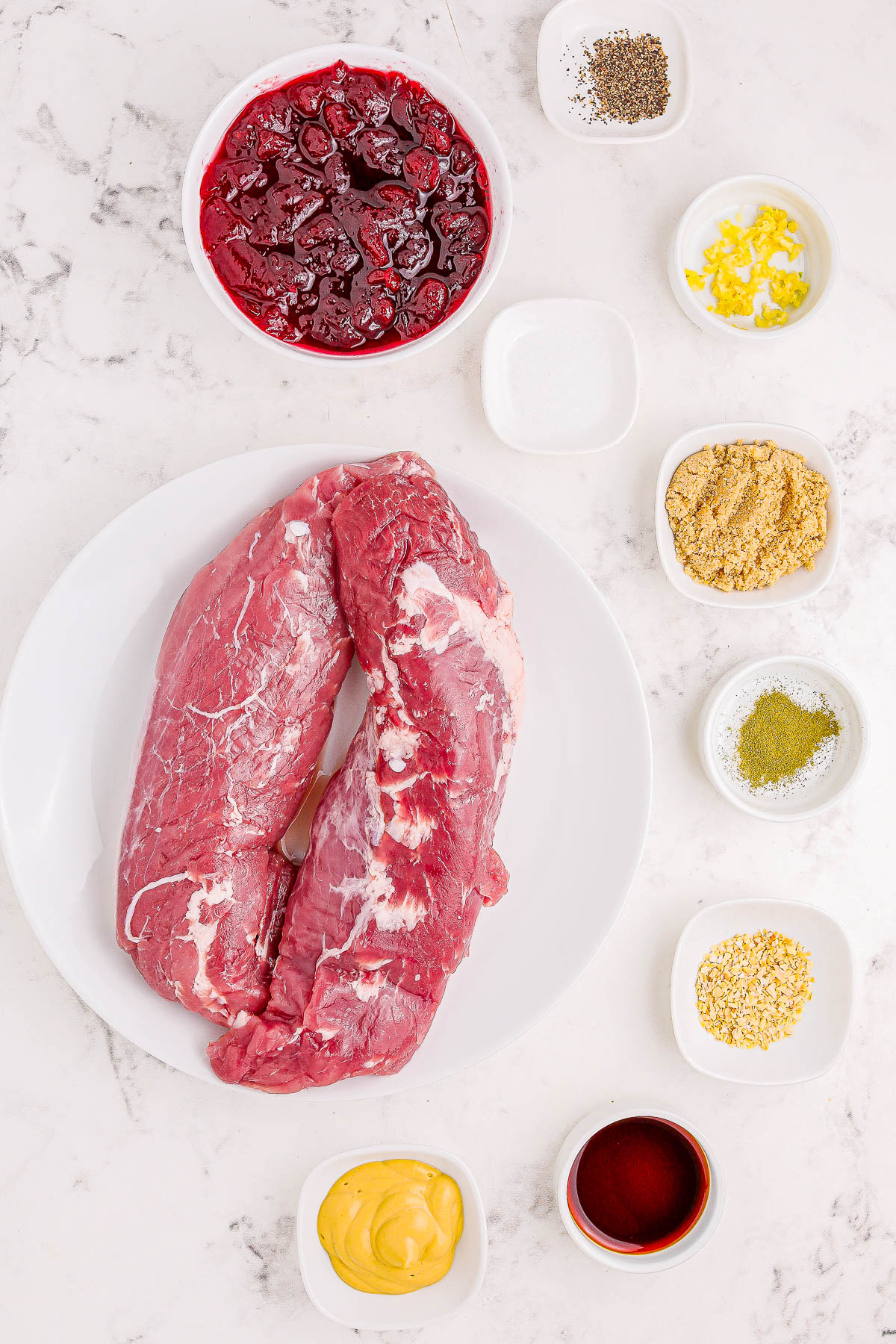 Raw beef tenderloin on a plate, surrounded by small bowls of mustard, red sauce, dry seasonings, lemon zest, and pepper, on a marble countertop.