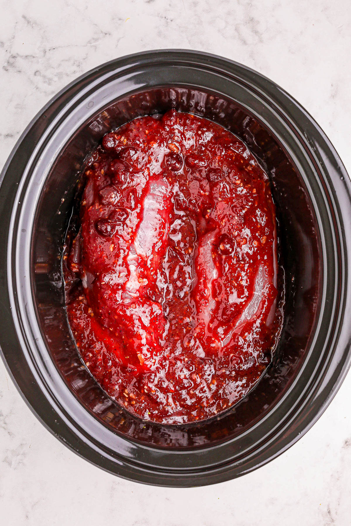 A meatloaf covered in red sauce in a black slow cooker on a marble surface.