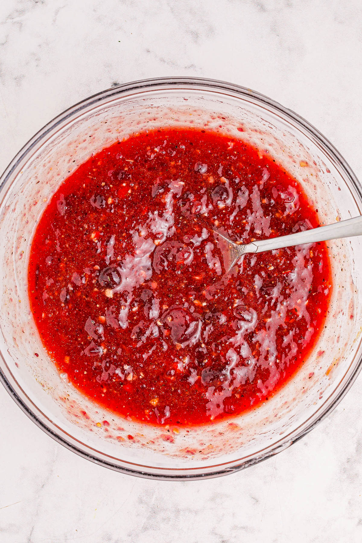 A clear bowl filled with a bright red, liquid mixture, possibly a sauce or dressing, with a metal spoon resting inside. The surface appears textured with visible small bits.