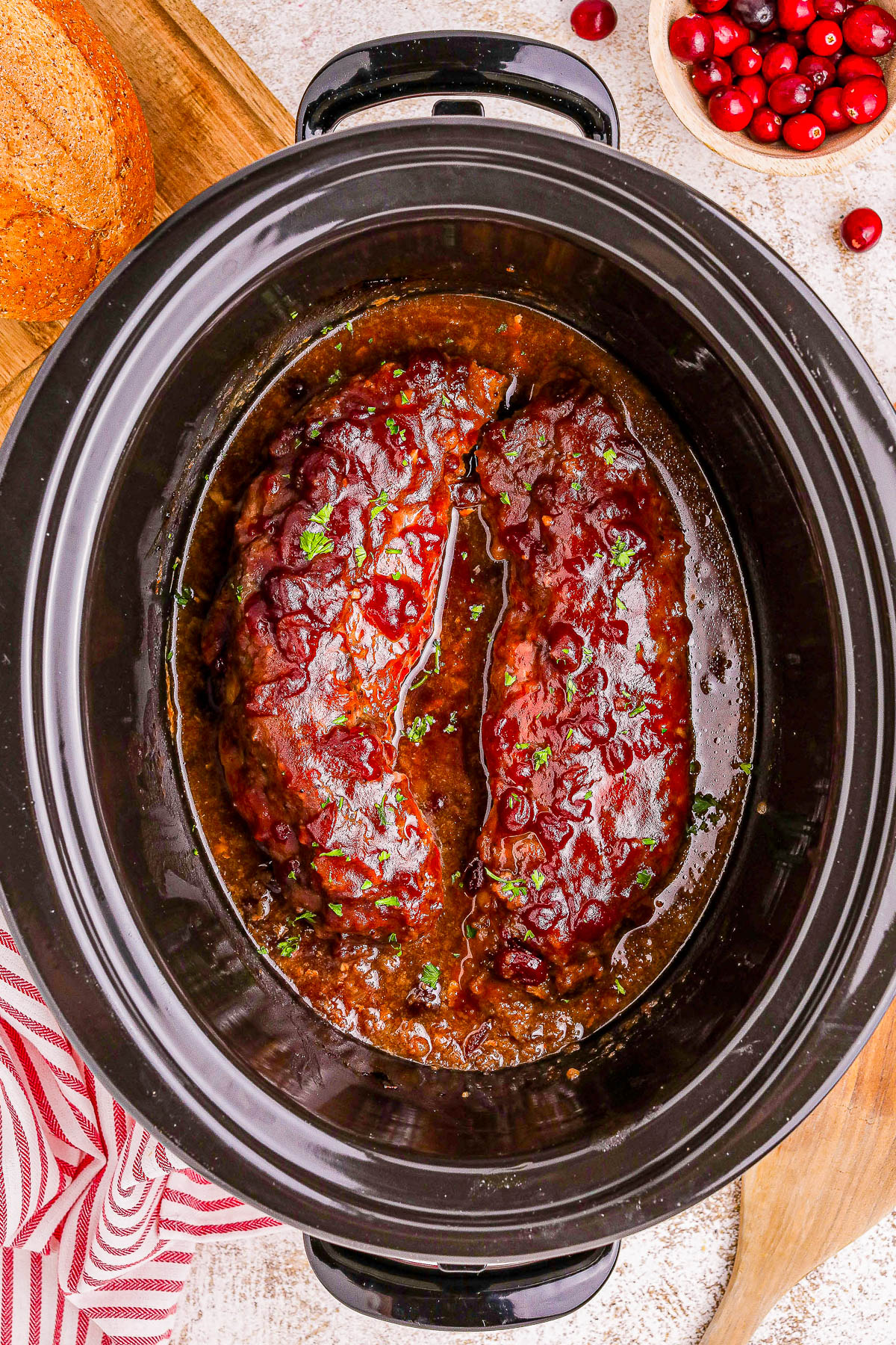 Two pork tenderloins in a slow cooker with a glaze, garnished with parsley. Cranberries and bread are nearby.