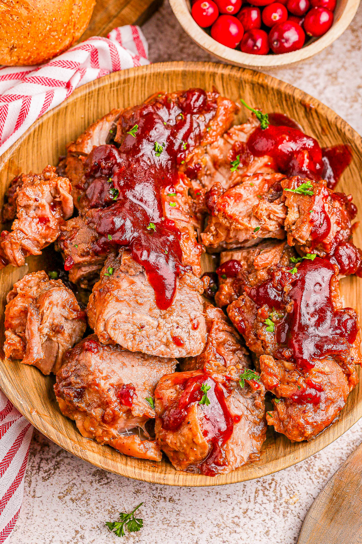 A wooden plate with sliced roast pork topped with cranberry sauce. Cranberries in a bowl are visible in the background.