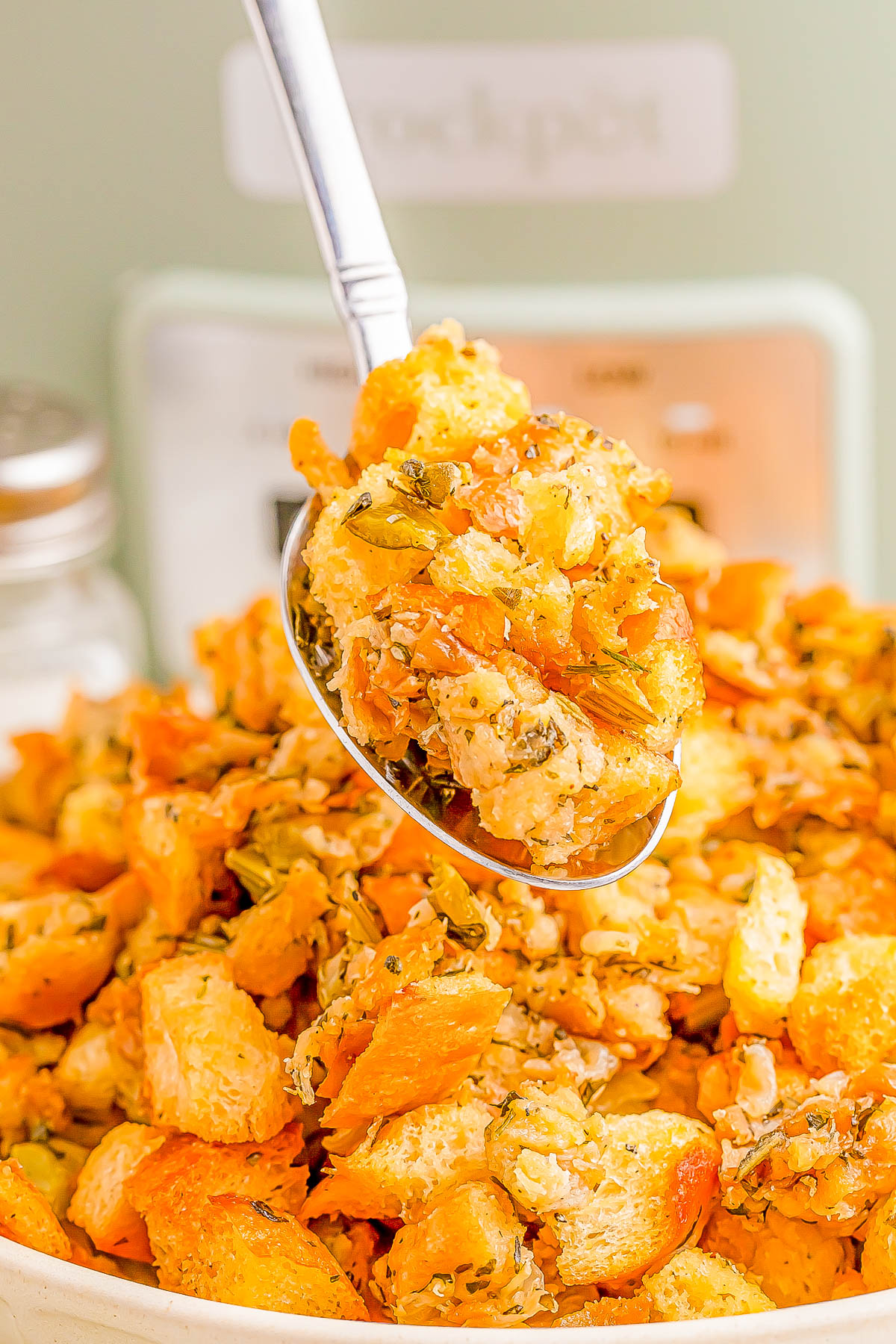 A spoonful of stuffing with bread cubes, carrots, and herbs, held above a bowl filled with more stuffing.