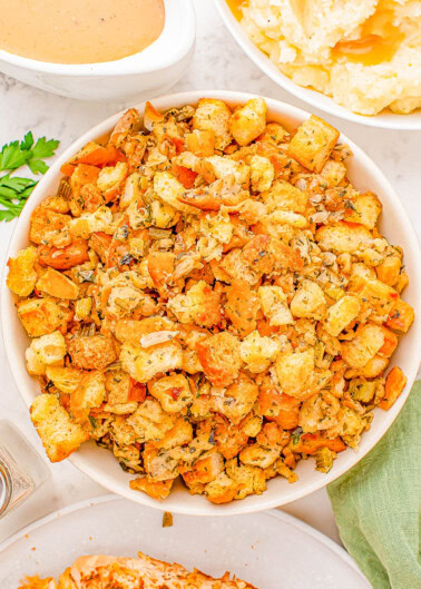 A bowl of stuffing made with croutons, herbs, and vegetables is placed on a table. Other dishes, including gravy and mashed potatoes, are partially visible nearby.