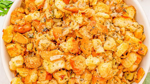 A bowl of stuffing made with croutons, herbs, and vegetables is placed on a table. Other dishes, including gravy and mashed potatoes, are partially visible nearby.