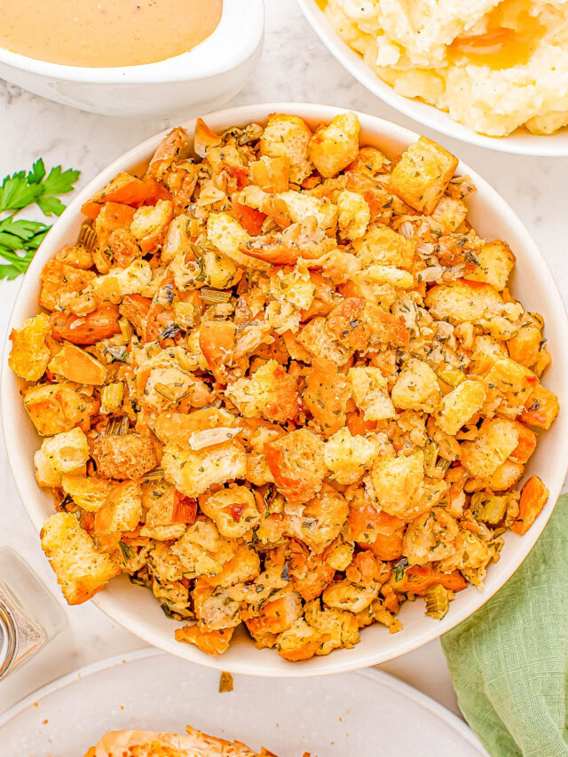 A bowl of stuffing made with croutons, herbs, and vegetables is placed on a table. Other dishes, including gravy and mashed potatoes, are partially visible nearby.