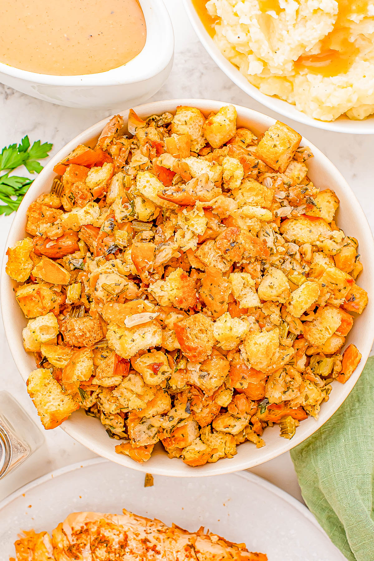 A bowl of stuffing made with croutons, herbs, and vegetables is placed on a table. Other dishes, including gravy and mashed potatoes, are partially visible nearby.