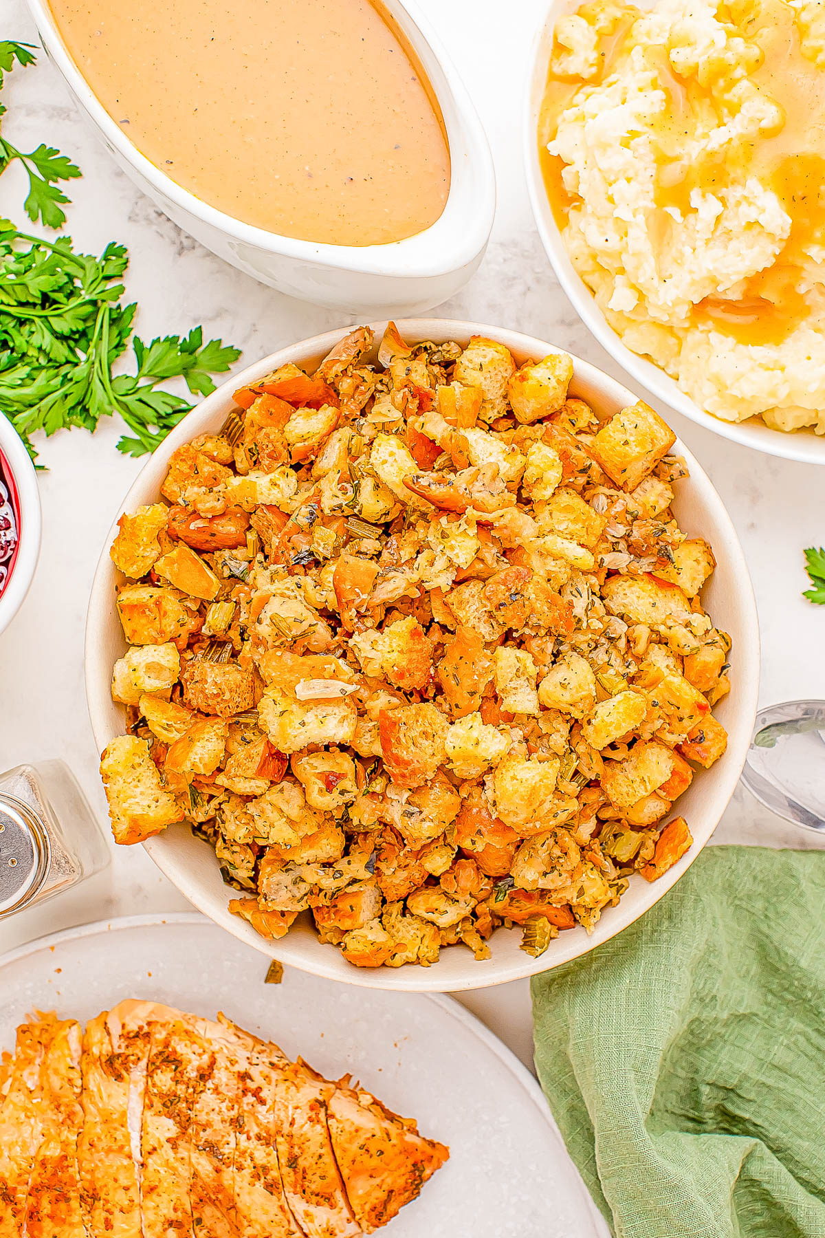 Bowl of stuffing surrounded by gravy, mashed potatoes, sliced turkey, and parsley.