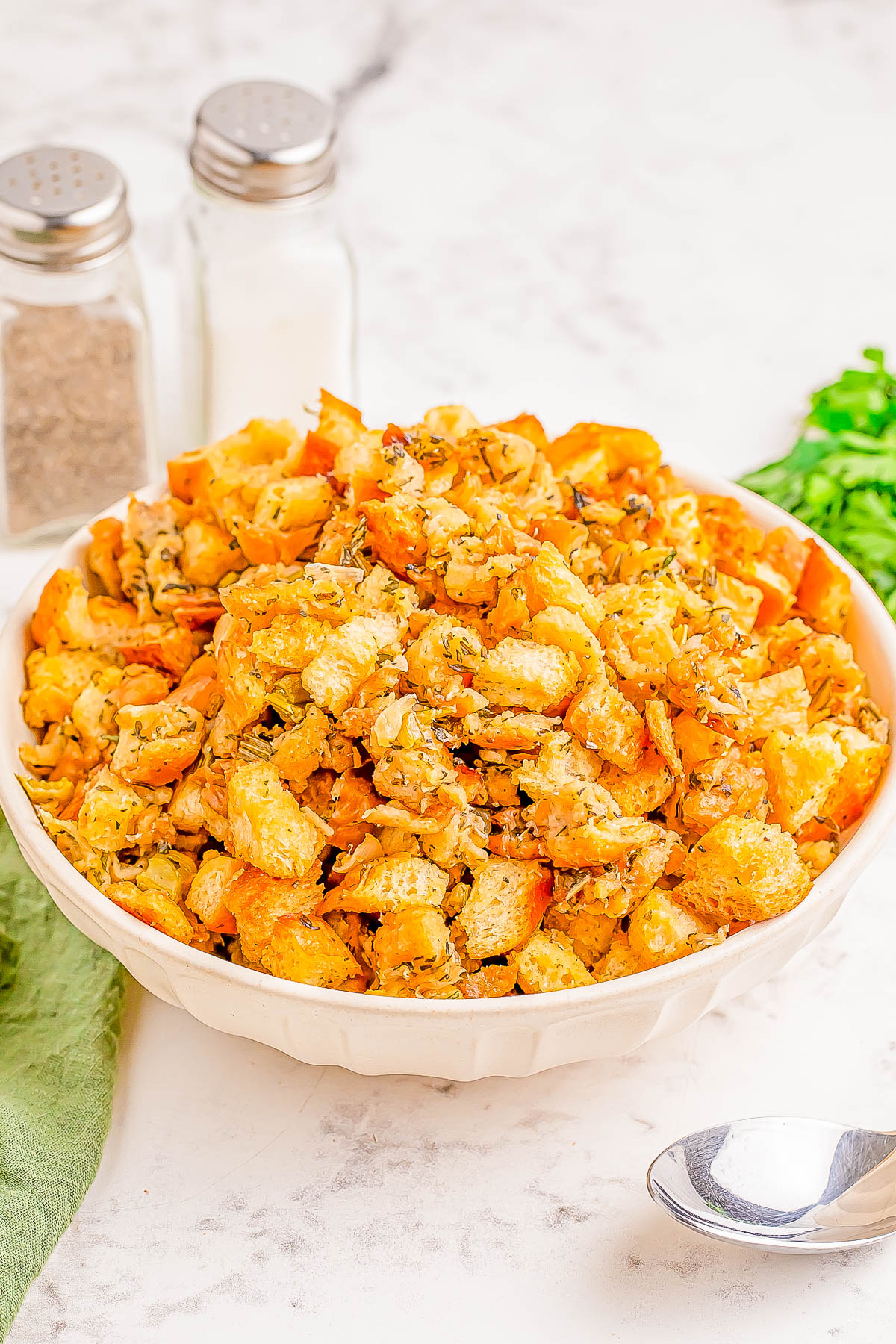 A bowl of seasoned bread stuffing on a marble countertop, with salt and pepper shakers, a green cloth, and a spoon nearby.