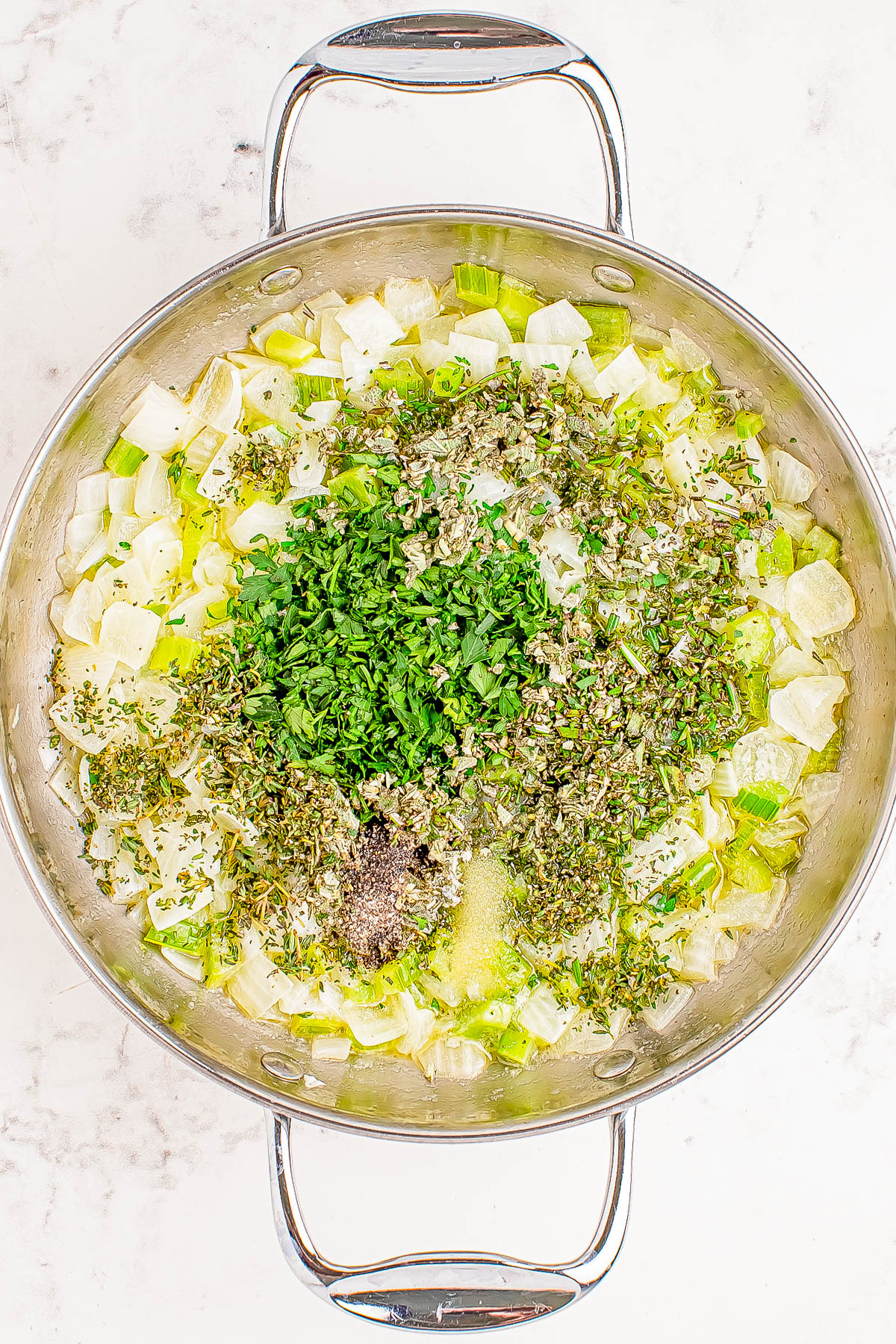 A large pot filled with diced onions, leeks, and a mix of fresh and dried herbs on a white background.