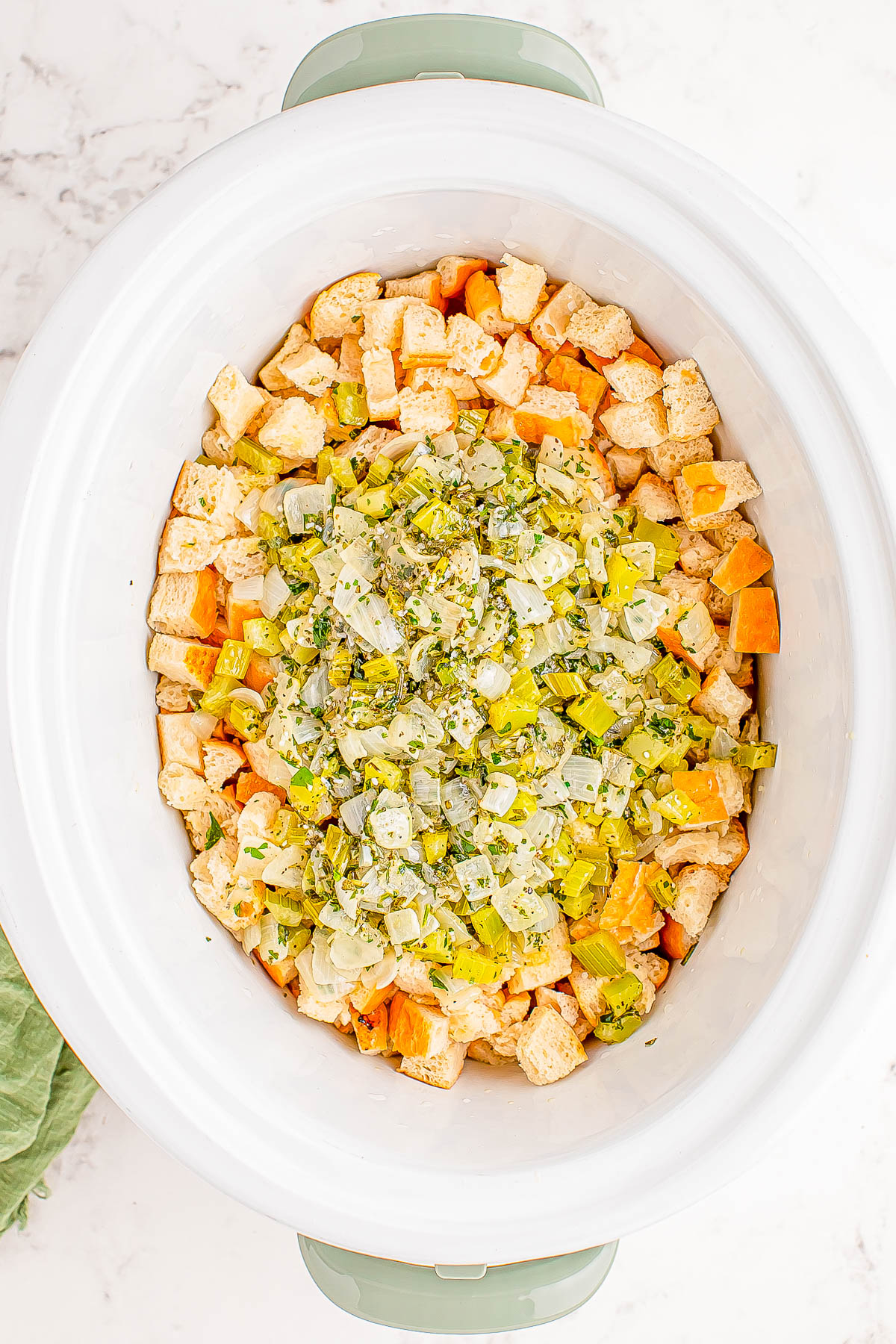 A white slow cooker filled with cubed bread, diced vegetables, and herbs ready for cooking.