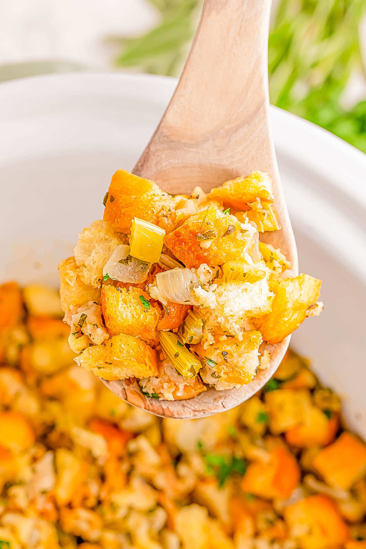 A wooden spoon holding a serving of stuffing with cubes of bread, diced vegetables, and herbs over a dish filled with more stuffing.