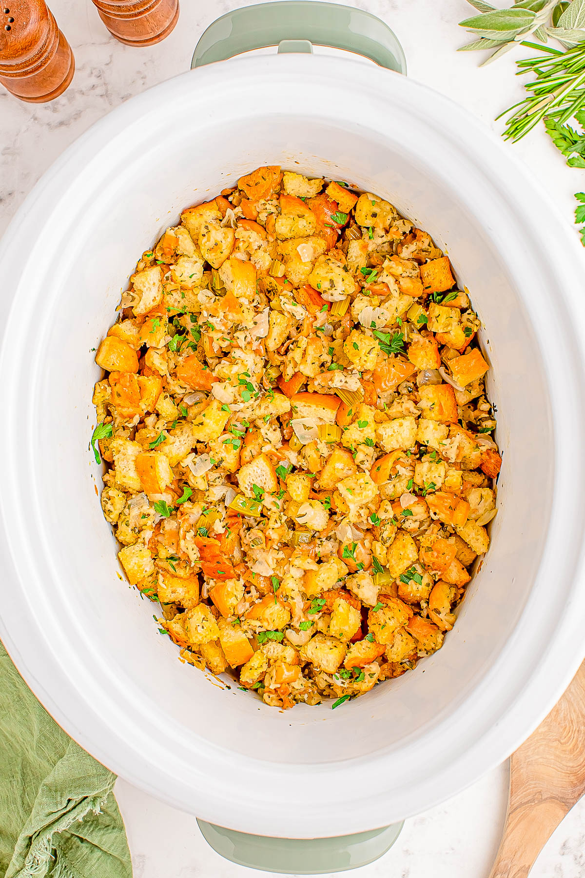 A slow cooker filled with a mixture of cubed bread, herbs, and vegetables, topped with chopped parsley.