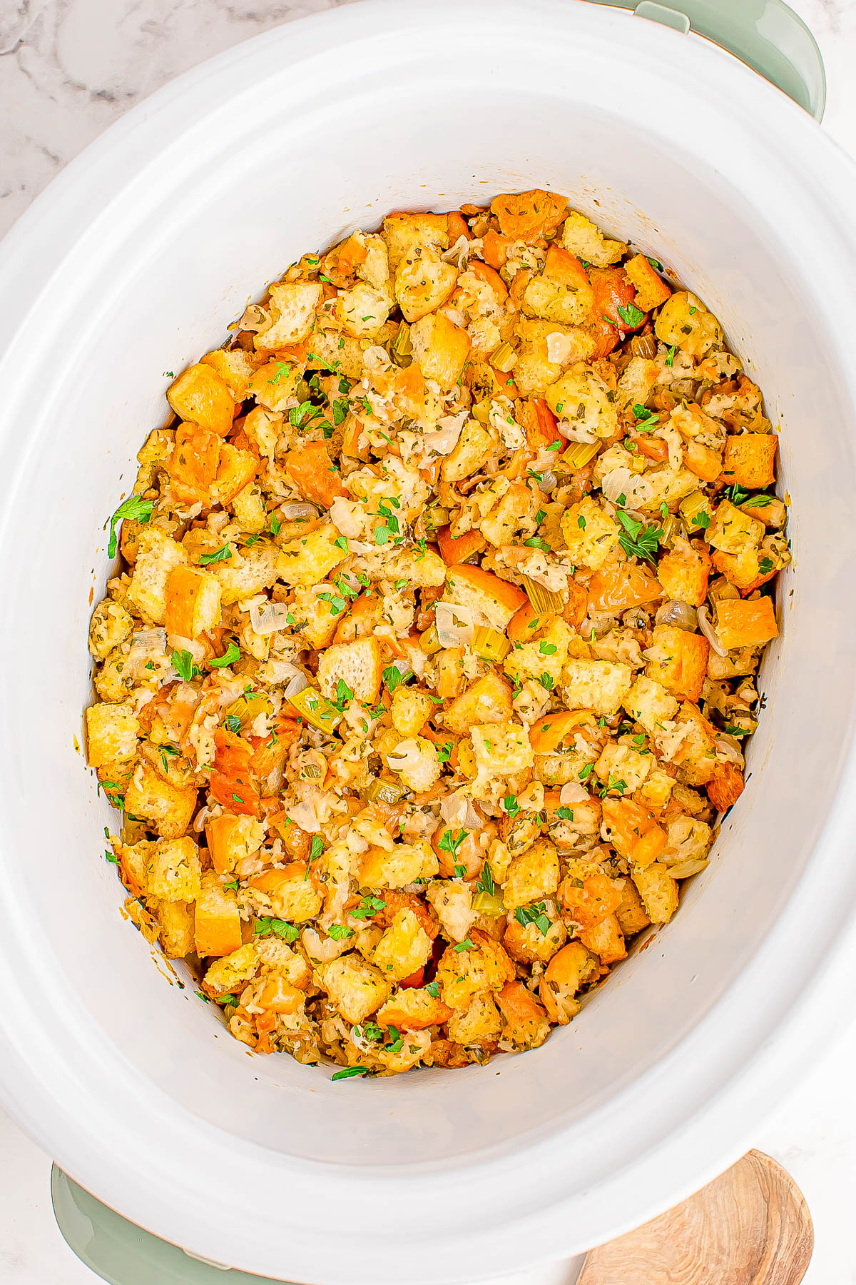 A white crock pot filled with cooked stuffing, featuring bread cubes, diced vegetables, and garnished herbs.