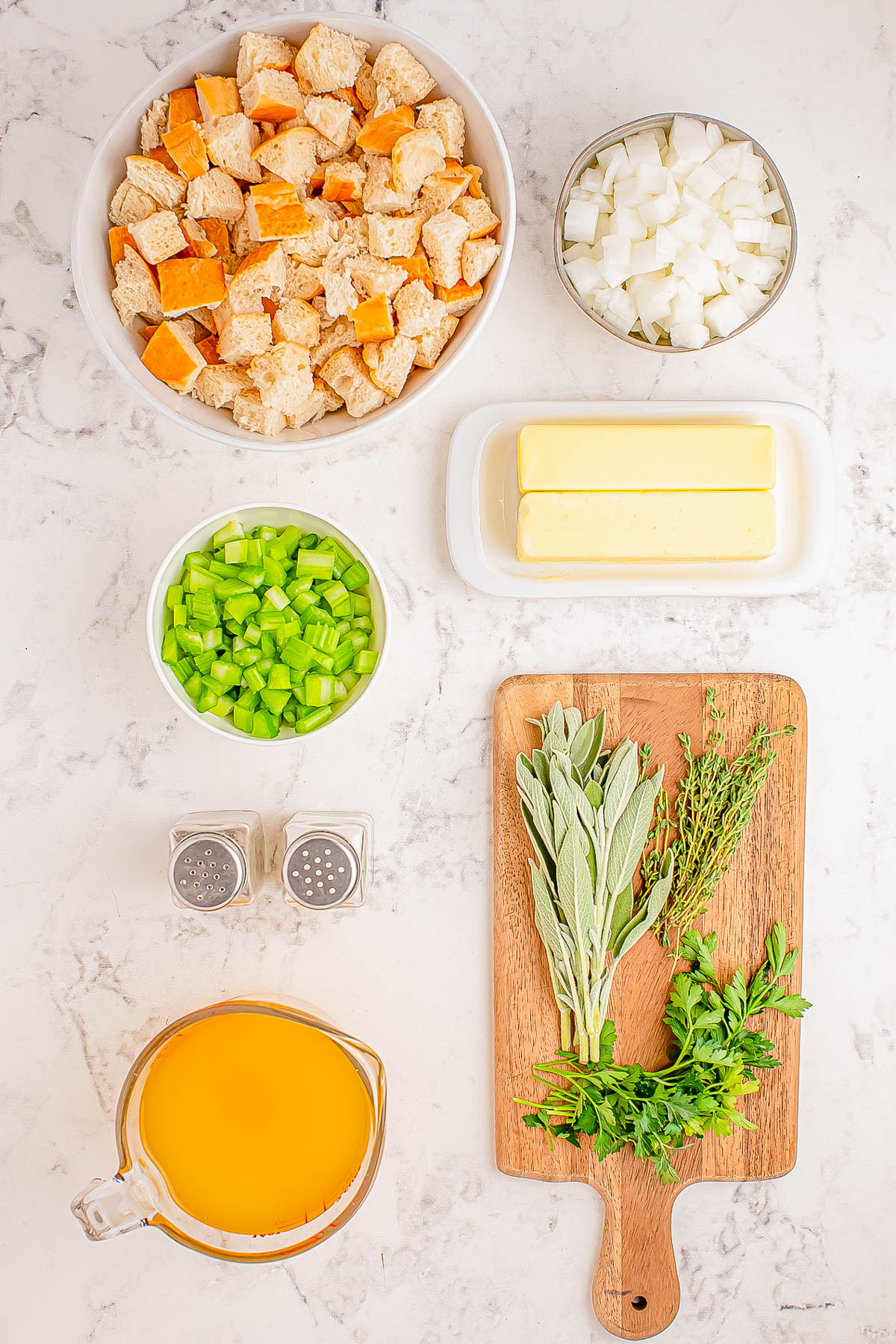 Ingredients displayed on a countertop: cubed bread, chopped onion, diced celery, butter sticks, fresh herbs on a board, salt and pepper shakers, and a measuring cup of broth.