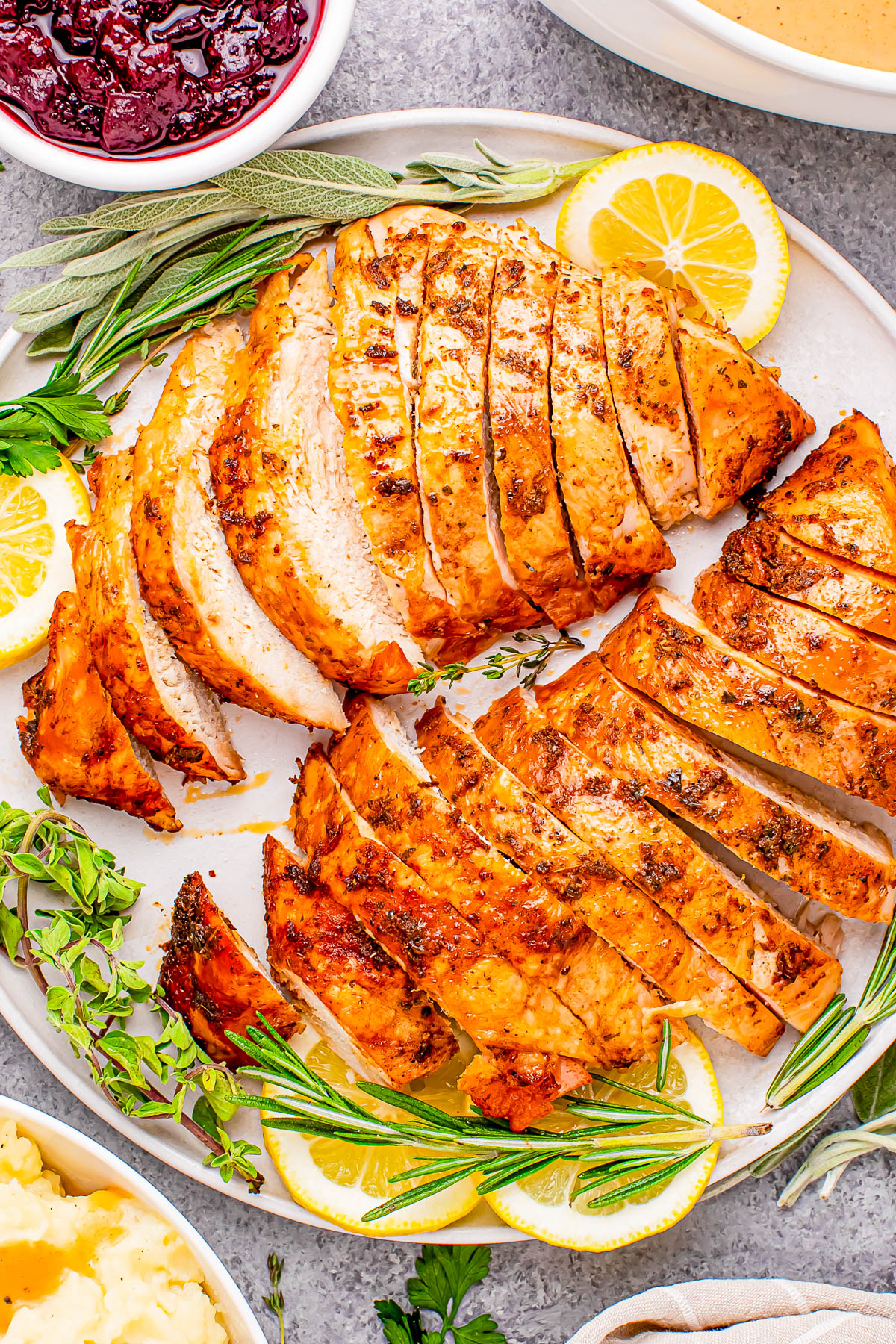 Sliced roasted turkey breast garnished with lemon slices and herbs, surrounded by sides like cranberry sauce and mashed potatoes.