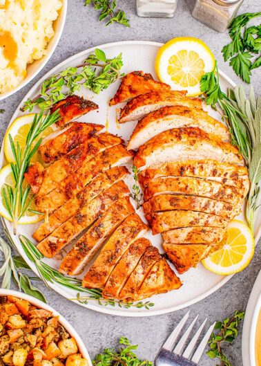 Sliced roasted turkey breast on a plate with lemon slices and herbs, surrounded by dishes of mashed potatoes and stuffing.