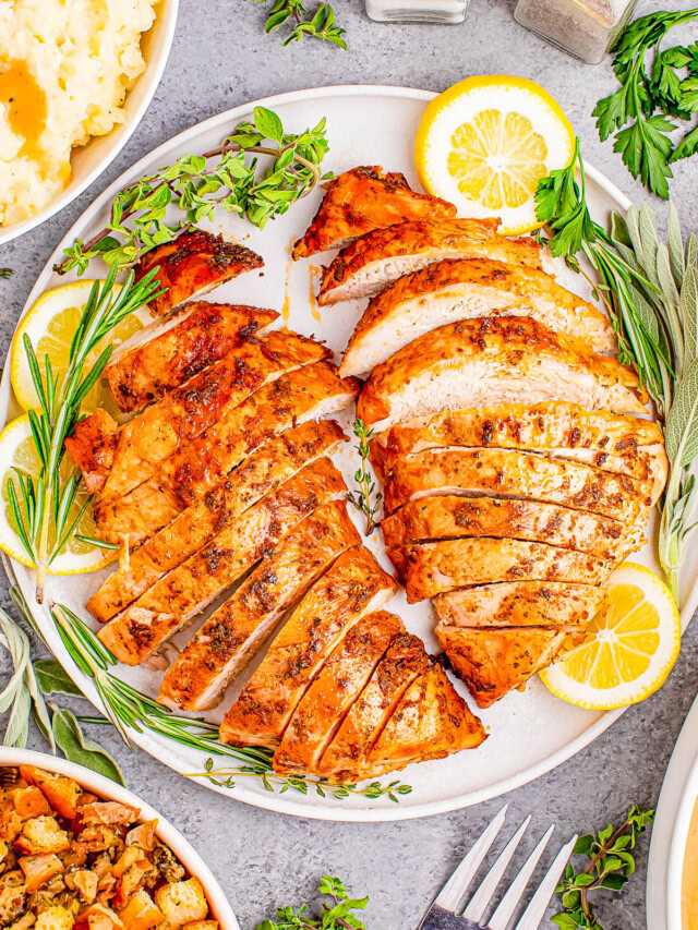 Sliced roasted turkey breast on a plate with lemon slices and herbs, surrounded by dishes of mashed potatoes and stuffing.