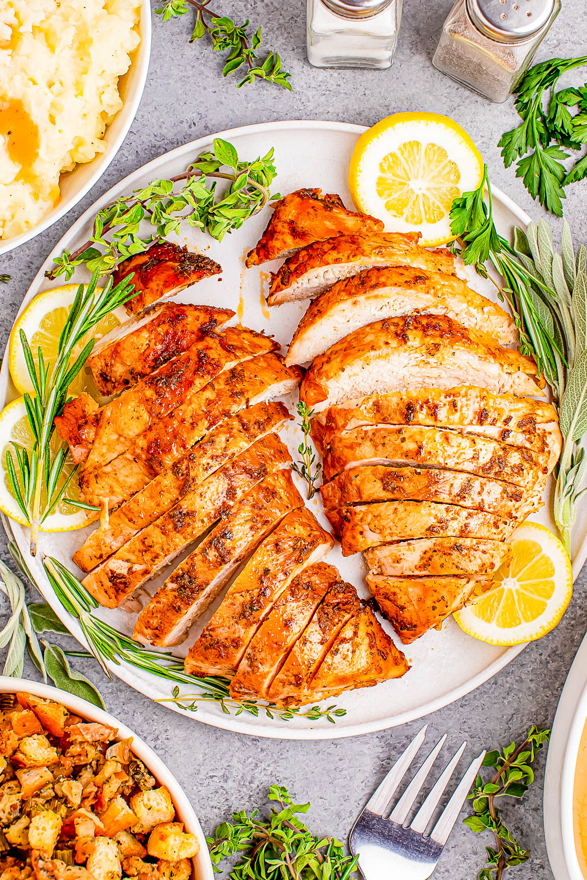 Sliced roasted turkey breast on a plate with lemon slices and herbs, surrounded by dishes of mashed potatoes and stuffing.