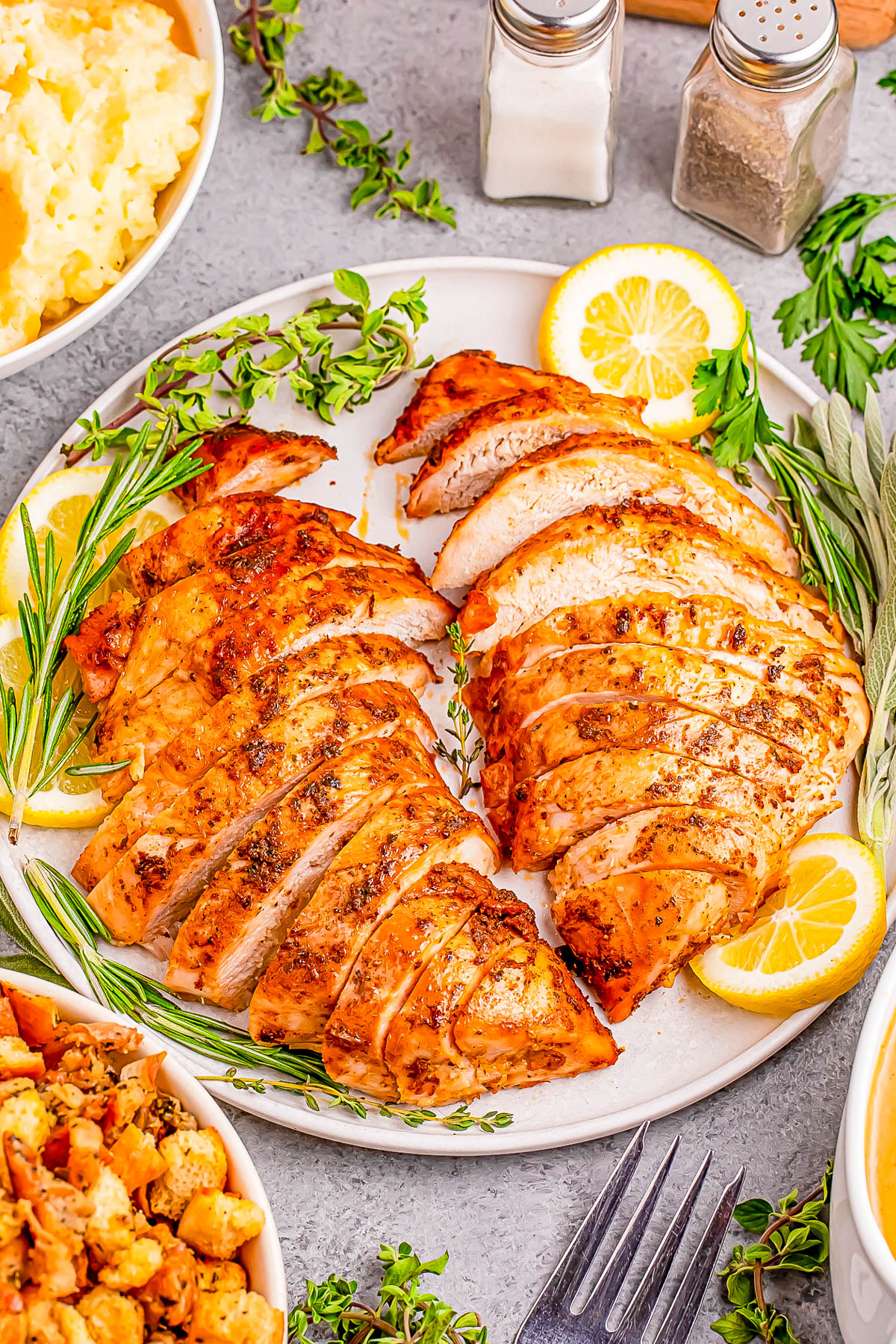 Sliced grilled turkey breast garnished with herbs and lemon slices, surrounded by mashed potatoes, stuffing, and seasoning shakers on a table.