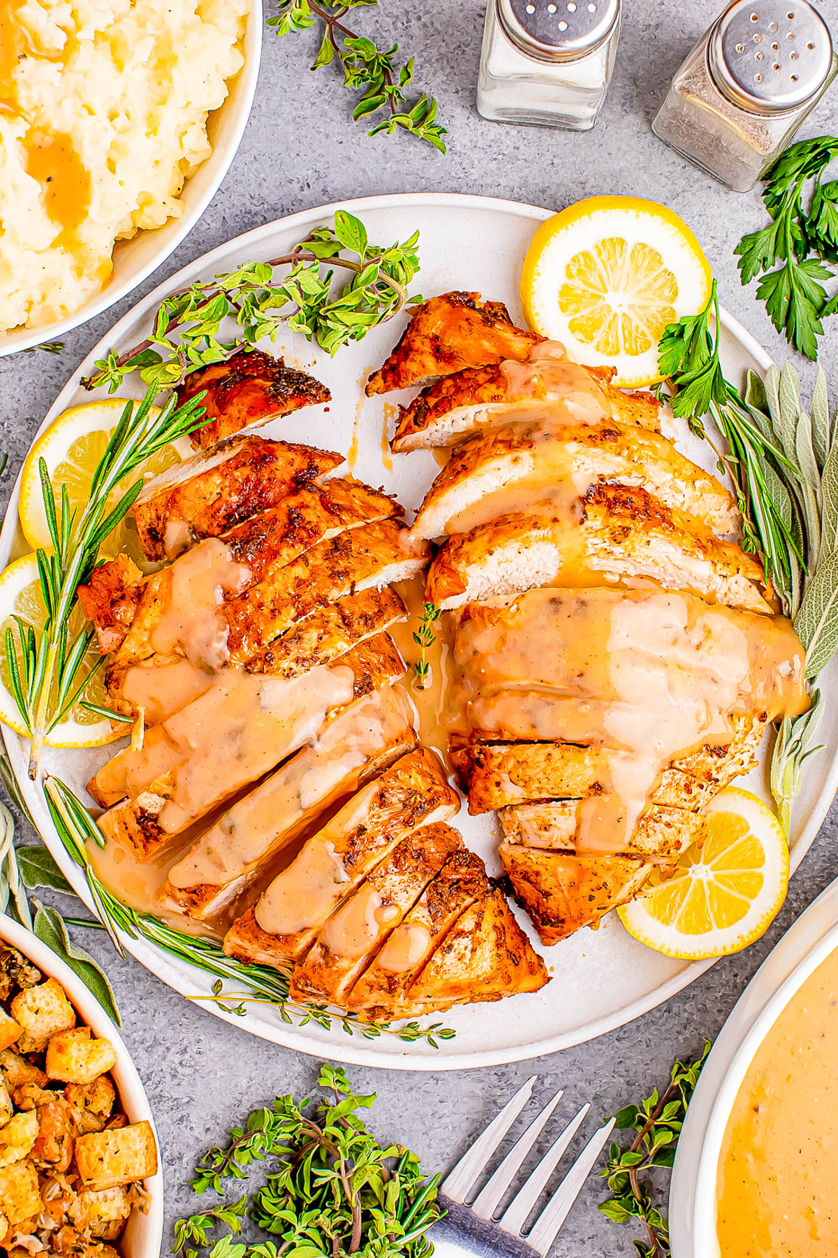 Sliced roasted chicken breast with gravy, garnished with lemon slices and herbs, served on a white plate. Mashed potatoes and stuffing visible on the side.