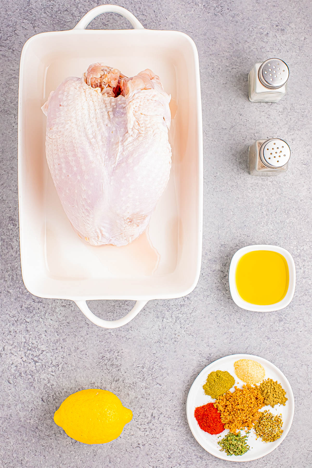 Raw turkey in a baking dish surrounded by spices, a lemon, oil in a bowl, and salt and pepper shakers on a gray countertop.