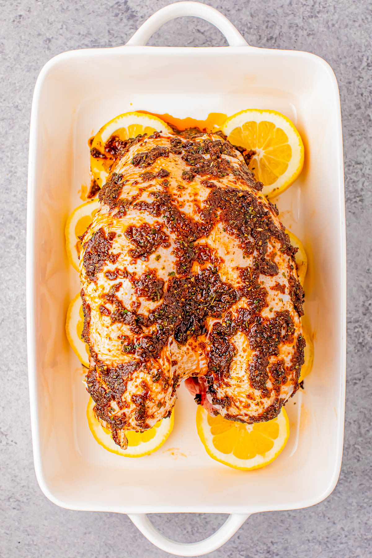 Roasted chicken in a white baking dish, seasoned with herbs and spices, surrounded by lemon slices.