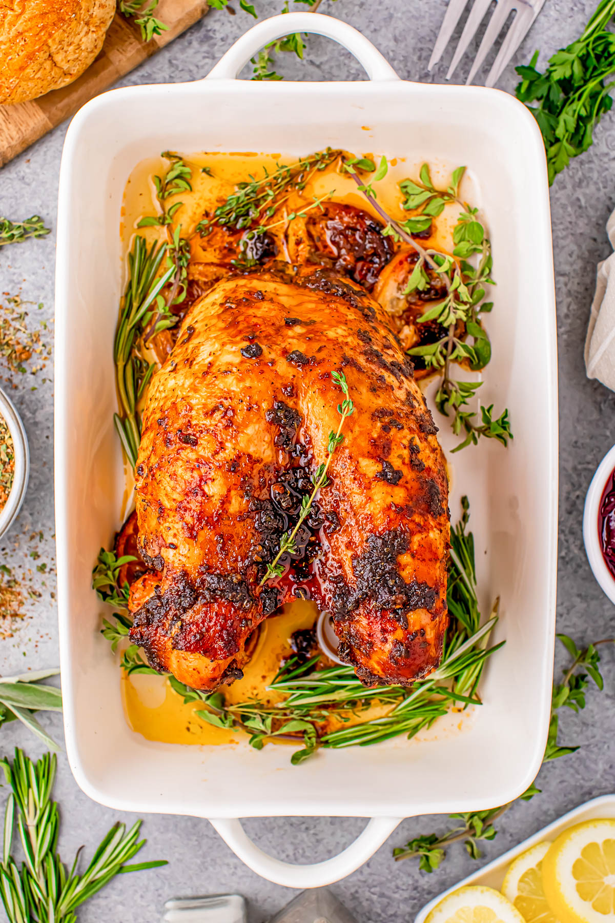 A roasted chicken seasoned with herbs like rosemary and thyme is placed in a white baking dish, surrounded by lemon slices and additional herbs.