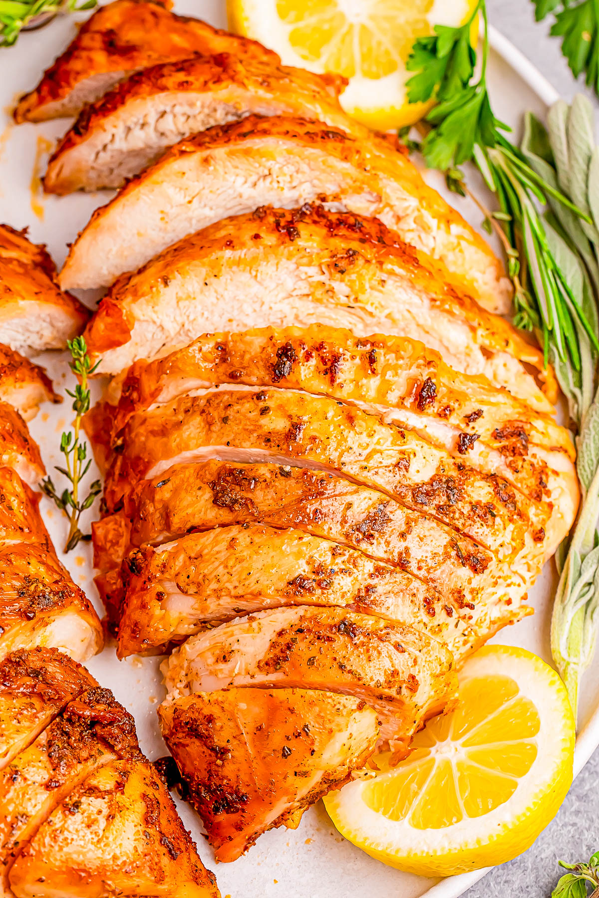 Sliced grilled chicken breast with seasoning, garnished with lemon slices, thyme, and sage leaves on a white plate.