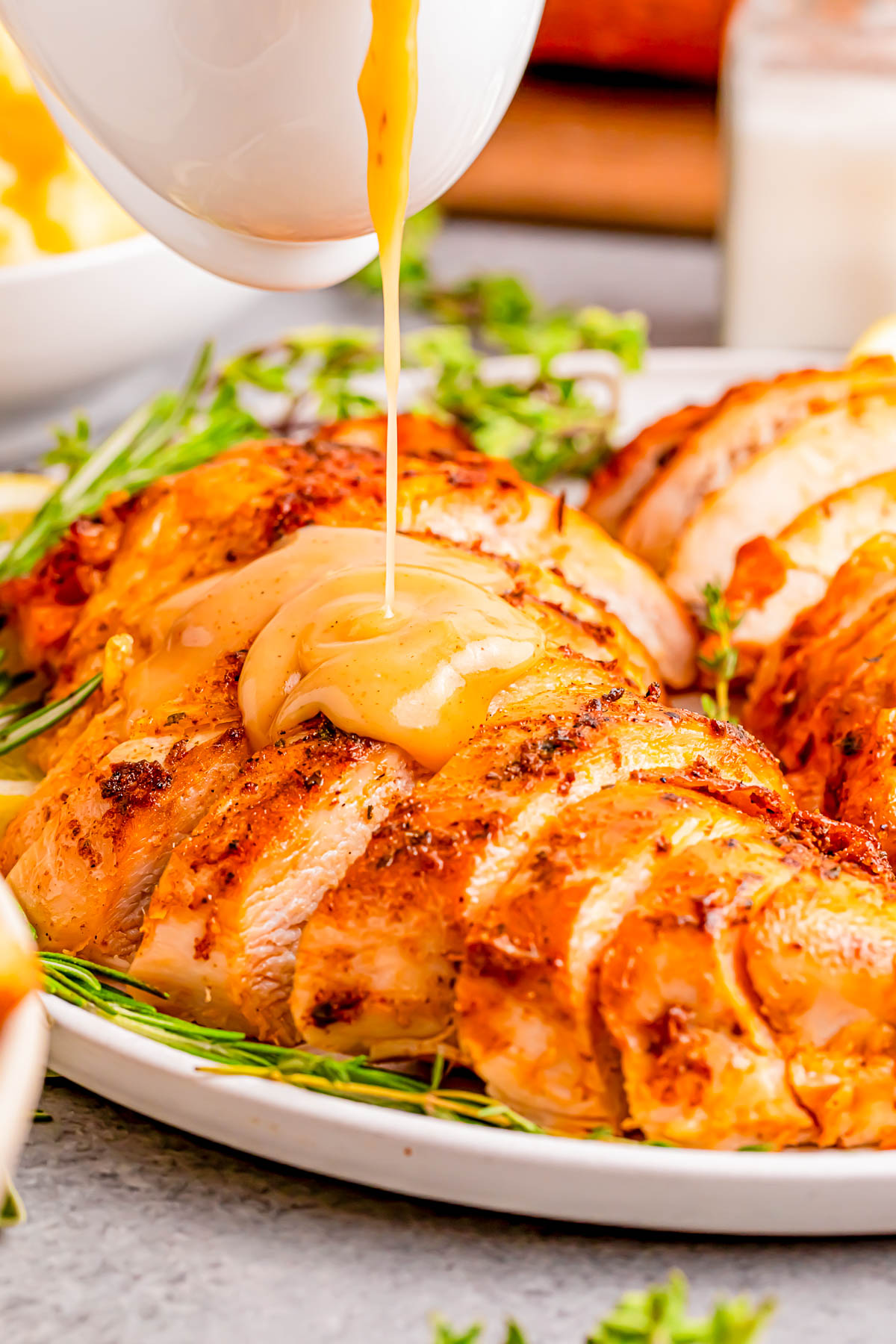 Sliced roasted chicken being drizzled with gravy on a white plate, garnished with herbs.