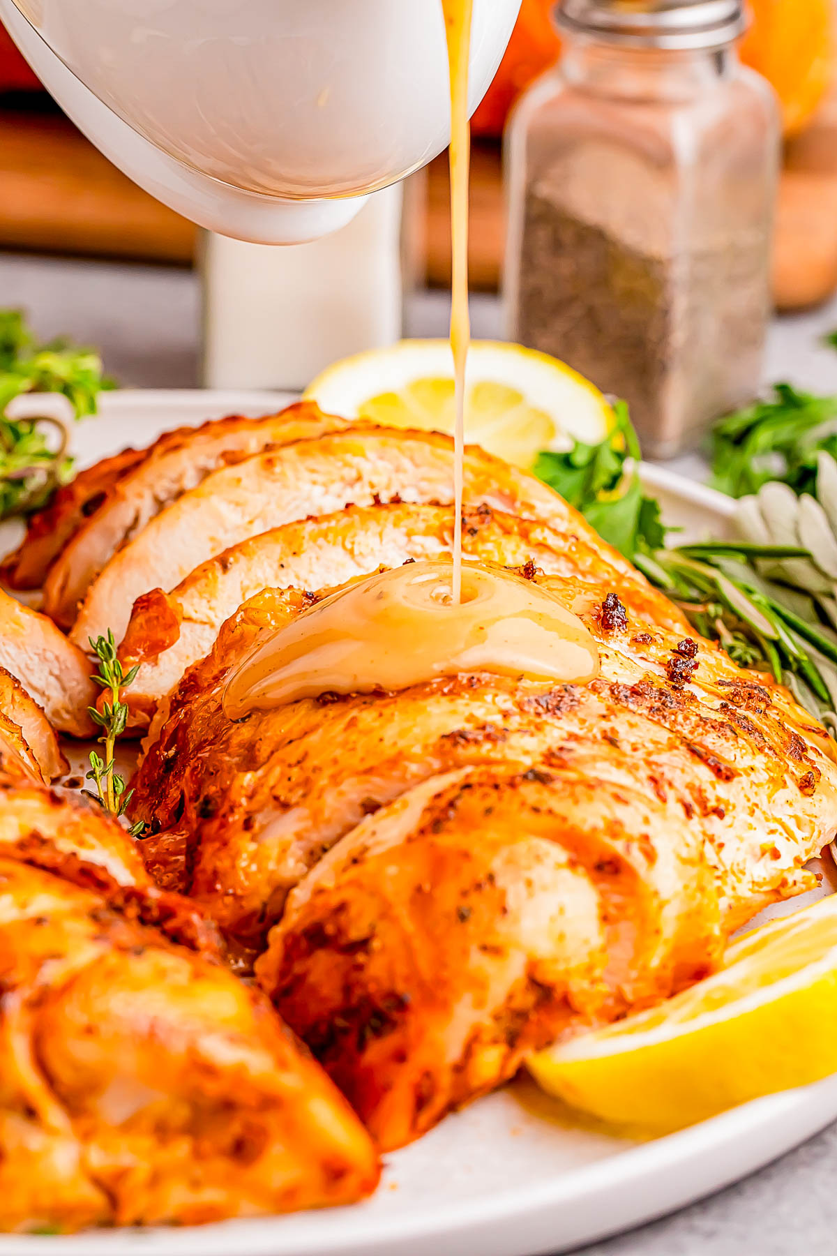 Sliced roasted chicken on a plate being drizzled with gravy, garnished with herbs and lemon wedges, with a salt shaker and herbs in the background.