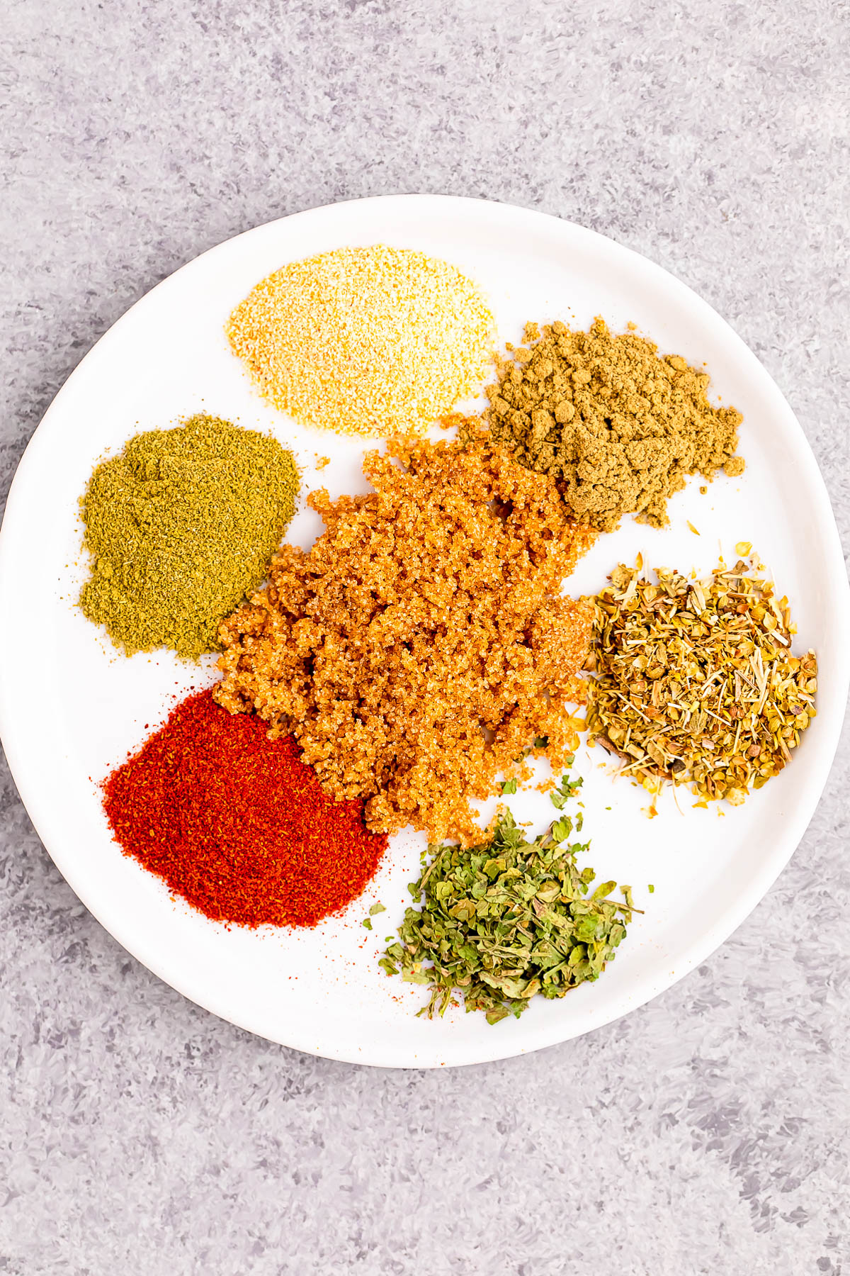 A white plate with seven different colorful spices and herbs arranged separately on a gray surface.