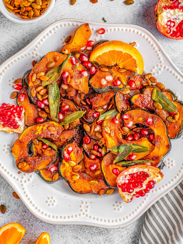 A plate of roasted acorn squash with pomegranate seeds, orange slices, and sage leaves, garnished with pumpkin seeds.